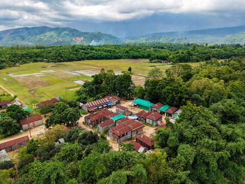 Aerial view of a rural village with corrugated metal roofs surrounded by lush green trees and agricultural fields near the mountains in Sunamganj, Sylhet Division, Bangladesh.