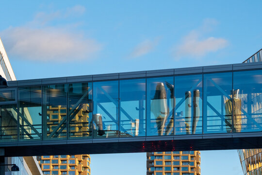 Two people appear as silhouette figures inside a glass skybridge connection over Stockholm, Sweden, where a blue office walkway crosses the open air