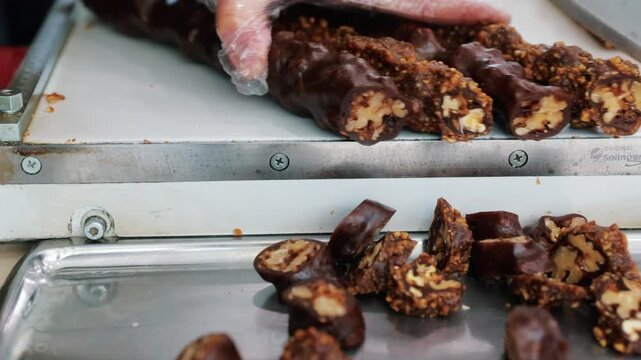 Professional close-up of traditional walnut sausage (cevizli sucuk) being sliced efficiently by an industrial cutter and falling onto a tray. Food production and Turkish cuisine concept.