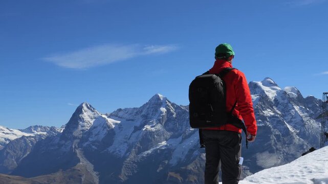Adventurous man with a backpack standing on a snowy peak and looking at the spectacular mountain range of the Swiss Alps in winter