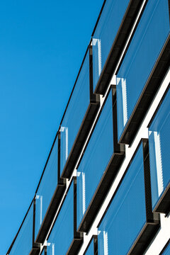 A glass panel facade catches blue sky reflection, where railing lines and windows create sharp urban geometry along the building edge