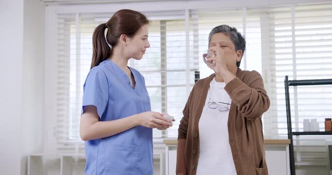 Slowmotion moment of Asian elderly woman drinking water after receiving medication from Caucasian young female physiotherapist in clinic during routine elderly rehabilitation support session