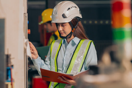 Group of diverse Asian technicians and female industry specialists collaborating next to production machinery, checking maintenance checklists and auditing plant operations to ensure workplace safety.