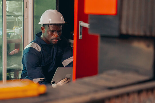 Black male maintenance specialist in safety gear tracking logistics data or automation software on a tablet screen during factory operation, showcasing digital transformation and workforce diversity.
