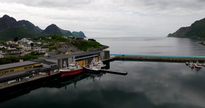 Hus&oslash;y harbor on Senja Island in Norway featuring fishing boats, fjord water, coastal village buildings, moody Nordic light and silhouetted mountain scenery, cinematic aerial view