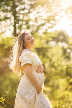 Pregnant woman enjoying golden sunlight in a floral dress outdoors, expecting mother smiling in summer