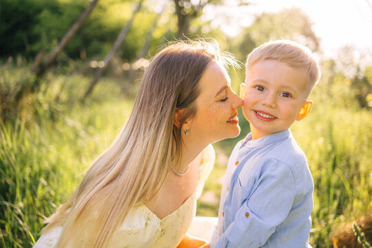 Mother kissing smiling son in sunlit meadow enjoying family love and outdoor summer warmth