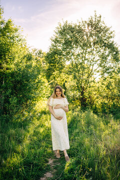 Pregnant woman in white dress smiling in a sunlit meadow during a maternity photoshoot