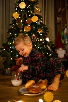 Little boy in plaid pajamas enjoying milk and doughnuts by a decorated Christmas tree at home