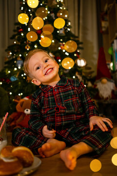 Smiling toddler in plaid pajamas sitting by a decorated Christmas tree with warm bokeh lights