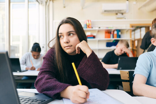 Thoughtful female student looking away while sitting at desk with book and laptop in classroom