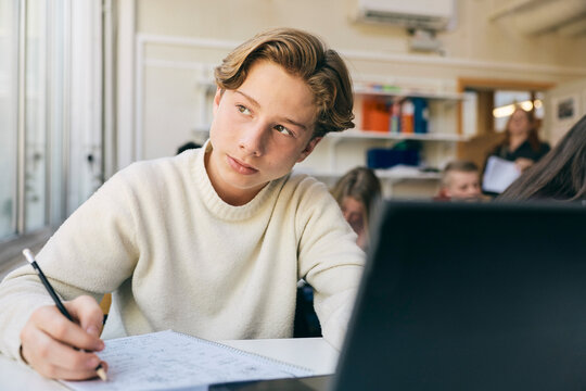 Thoughtful teenage boy looking away while solving math equation in classroom at desk