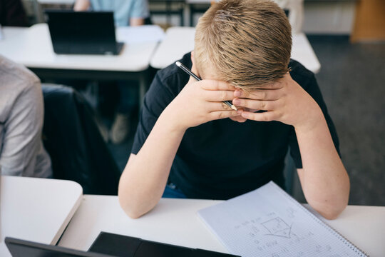 High angle view of teenage boy sitting with head in hands at desk while studying in classroom