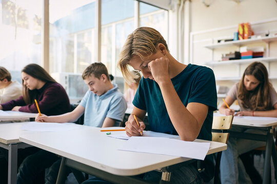 Focused teenage schoolboy writing exam paper while sitting at desk in classroom