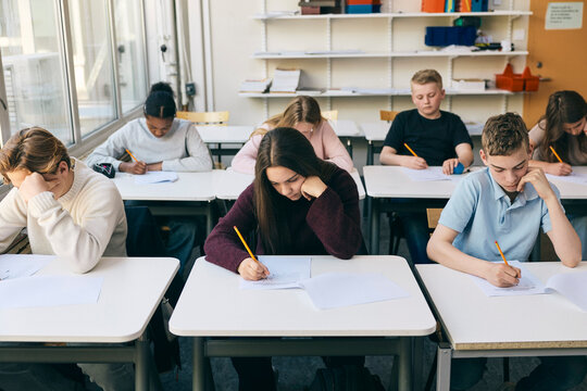 Teenage girl writing educational exam while sitting with classmates at desk in junior high school
