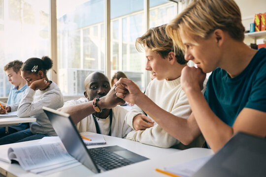 Male teacher giving fist bump to teenage boy studying in classroom with laptop at school