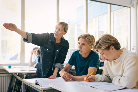 Female teacher gesturing while explaining teenage boys sitting at desk in classroom at junior high school