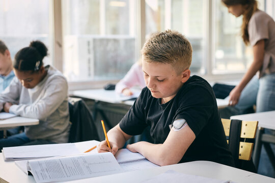 Male student wearing glucose monitor on arm writing in book while studying at school
