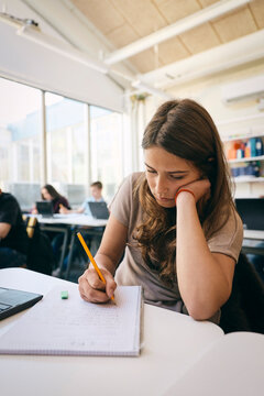 Teenage girl writing in book while leaning on elbow at classroom desk