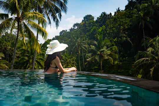 Woman wearing a hat relaxing in an infinity pool with views of lush jungle and rice terraces in Ubud, Bali, Indonesia.