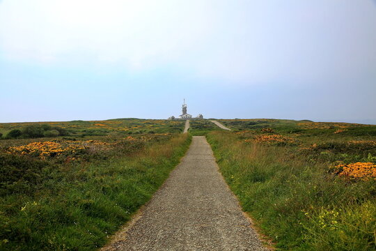 Ein Schotterweg f&uuml;hrt durch die flache, gr&uuml;n-gelbe Heidelandschaft direkt auf die historische Signalstation (S&eacute;maphore) an der zerkl&uuml;fteten Atlantikk&uuml;ste der Bretagne.