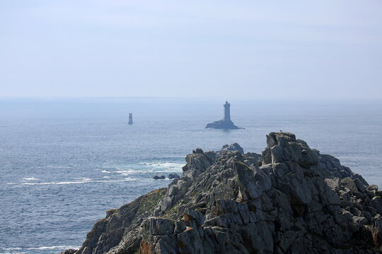 Raue Felsklippen fallen steil in den Atlantik ab, w&auml;hrend im Hintergrund die ber&uuml;hmten Leuchtt&uuml;rme Phare de la Vieille und T&eacute;vennec aus den unruhigen Wellen ragen.