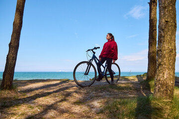 Fototapeta premium Young woman in red jacket standing with bicycle near trees by seaside under clear blue sky