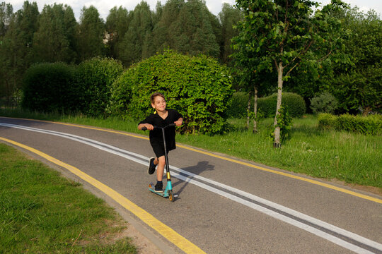 Happy active boy riding fast kick scooter on asphalt road in summer park, profile view of child driving, fast speed motion, childhood outdoor activity, healthy lifestyle concept, copy space