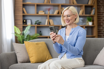 Obraz premium Blonde middle-aged woman in glasses sitting on a cozy living room couch, smiling and relaxed while browsing and messaging on her smartphone, enjoying digital connection and leisure