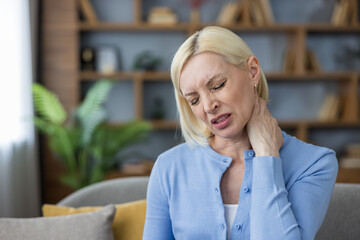 Fototapeta premium Middle-aged woman sitting on a sofa at home, feeling aching pain in her neck and shoulder, a concept representing chronic discomfort, stress, or muscle stiffness