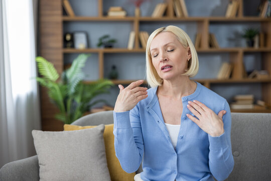Blonde middle-aged woman on a sofa fanning herself during a sudden hot flash at home, flushed and sweating from menopause-related discomfort and hormonal imbalance