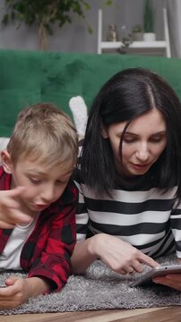 Family concept where attractive contented cheerful mother and her two 8-10s sons which lying on fluffy carpet and previsioning modern apps on tablet pc