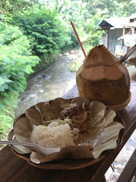 Traditional grilled chicken, rice, and fresh coconut drink served beside a flowing river in a lush tropical environment, ideal for travel, food, and lifestyle concepts.