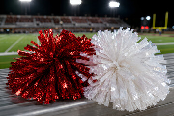 Red and white cheerleading pompoms lying on stadium bench at night, close-up with football field background