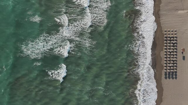 Aerial view of ocean waves rolling onto sandy beach with rows of empty umbrellas