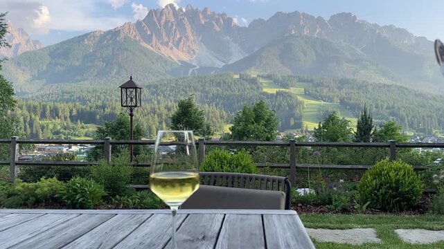 Elegant lifestyle footage of two wine glasses set against the stunning panoramic mountain views of the Dolomites, South Tyrol &mdash; a hand reaches in to take a glass in this cinematic travel moment.