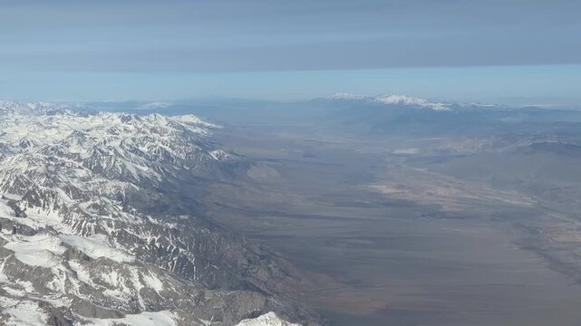 A wide aerial shot follows the spine of a heavily snow-laden mountain range &mdash; the jagged peaks and deep couloirs dusted brilliant white &mdash; as the terrain drops sharply into an enormous flat desert basi