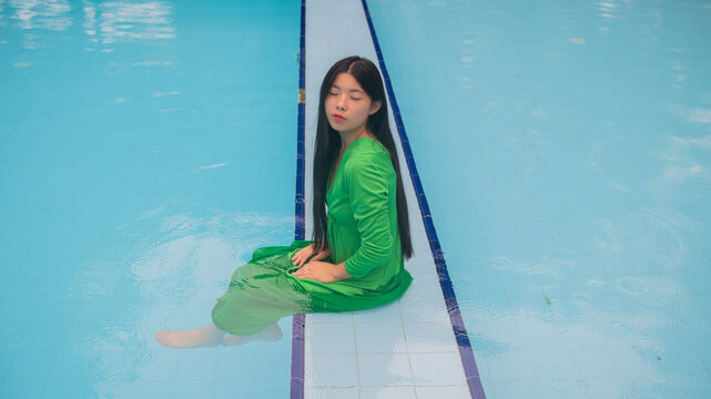 art portrait of young asian woman in green dress sitting on edge of swimming pool
