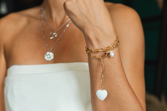 Close up of a woman hand wearing a gold chain bracelet with a white heart charm in front of layered silver necklaces.