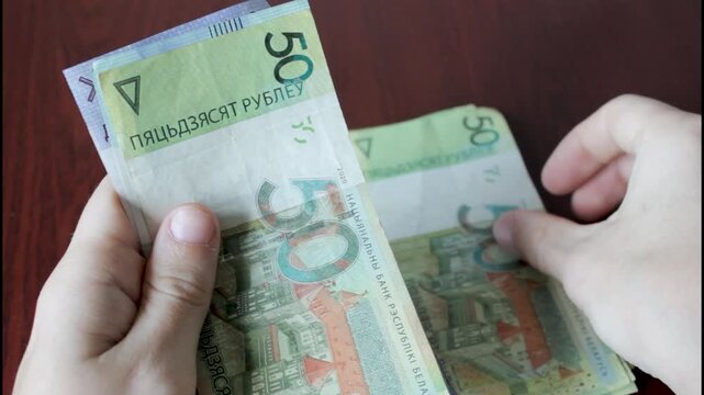 
A close-up of hands counting and stacking Belarusian banknotes on a wooden table. The banknotes are in denominations of 50, 100, and 200 BYN.
