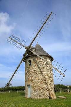 Historische Windm&uuml;hle Moulin de Trouguer aus Naturstein in der Bretagne unter blauem Himmel mit imposanten Holzfl&uuml;geln und einem liegenden M&uuml;hlstein im gr&uuml;nen Gras.