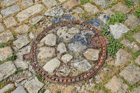 Ein runder, gusseiserner Schachtdeckel ist harmonisch mit Pflastersteinen ausgef&uuml;llt und f&uuml;gt sich unauff&auml;llig in das historische Kopfsteinpflaster einer Stra&szlig;e in Locronan ein.