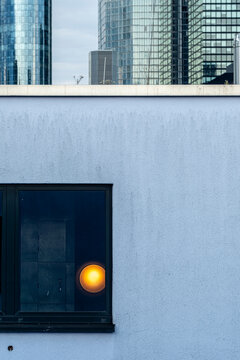 A single window lamp glows on a blue wall, while towers reflection and facade layers create urban architecture contrast around the frame