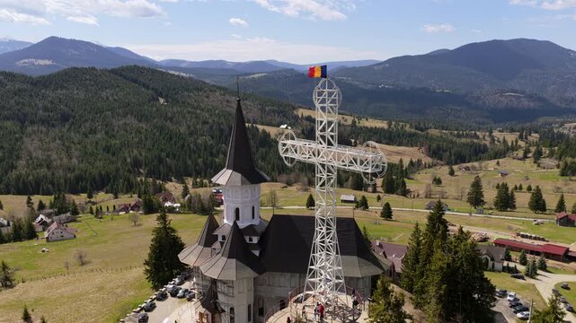 Aerial drone view of a Romanian flag waving above the large metal cross near Acoperamantul Maicii Domnului Monastery with the mountains in the background