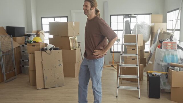 Man with arms crossed beside a ladder and stacked cardboard boxes in a building; contentment readiness.