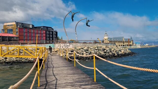 A man walks his dog from a wooden pier toward the Costanera promenade with the Monumento al Viento in Puerto Natales, Chile, on a sunny day along the waterfront