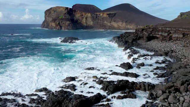A drone view approaching Capelinhos Volcano with waves crashing on dark lava rocks in the Azores