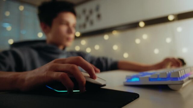 Close-up of a young gamer's hand on a computer mouse playing a video game at night. The keyboard and mouse have RGB lighting.