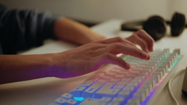 A teenage boy types on a backlit gaming keyboard while playing a video game or coding online at night. Close-up on his hands and focused face.