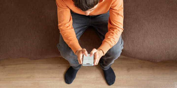 Teenager sitting on sofa and using smartphone viewed from above. Concept of screen time, social media, digital communication, online lifestyle and technology use among young people.